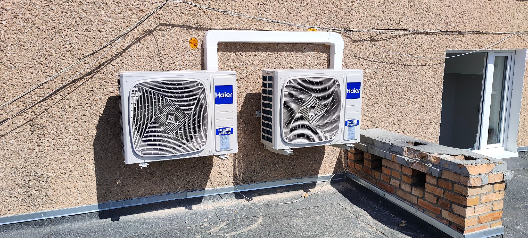 Dwa klimatyzatory Haier zamontowane na zewnątrz budynku, połączone białym przewodem, widoczne okno i fragment muru z cegły.