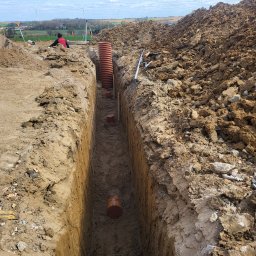 Przemysław Smektalski F.U. - Wykopany rów z widocznymi rurami kanalizacyjnymi, częściowo zakopanymi. W tle osoba i wiatraki.