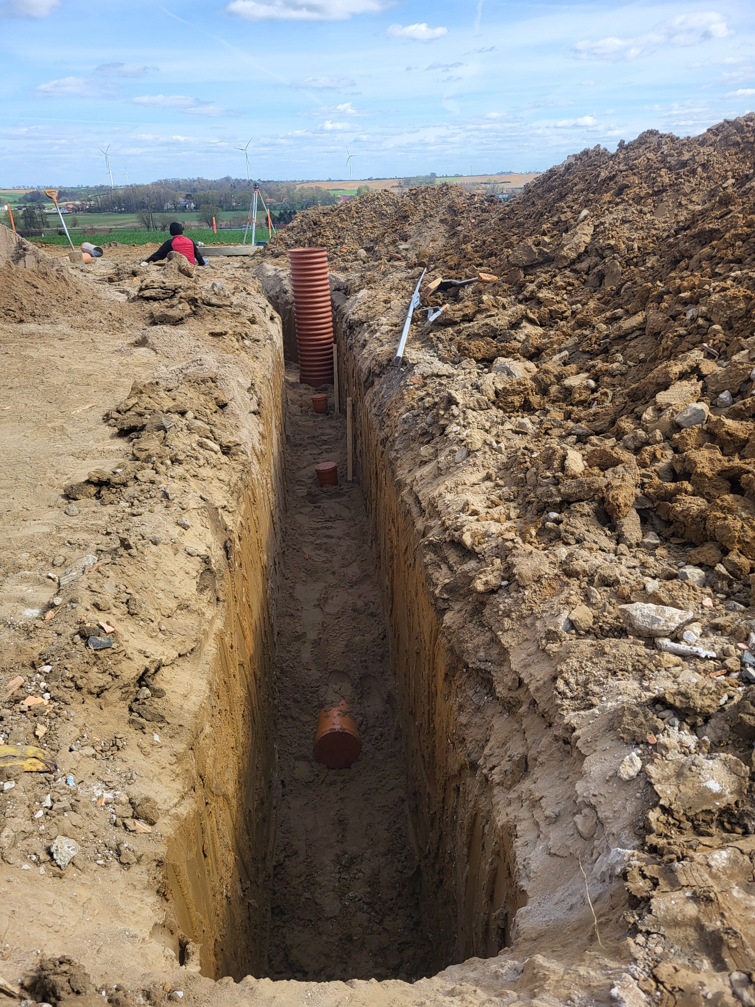 Wykopany rów z widocznymi rurami kanalizacyjnymi, częściowo zakopanymi. W tle osoba i wiatraki.