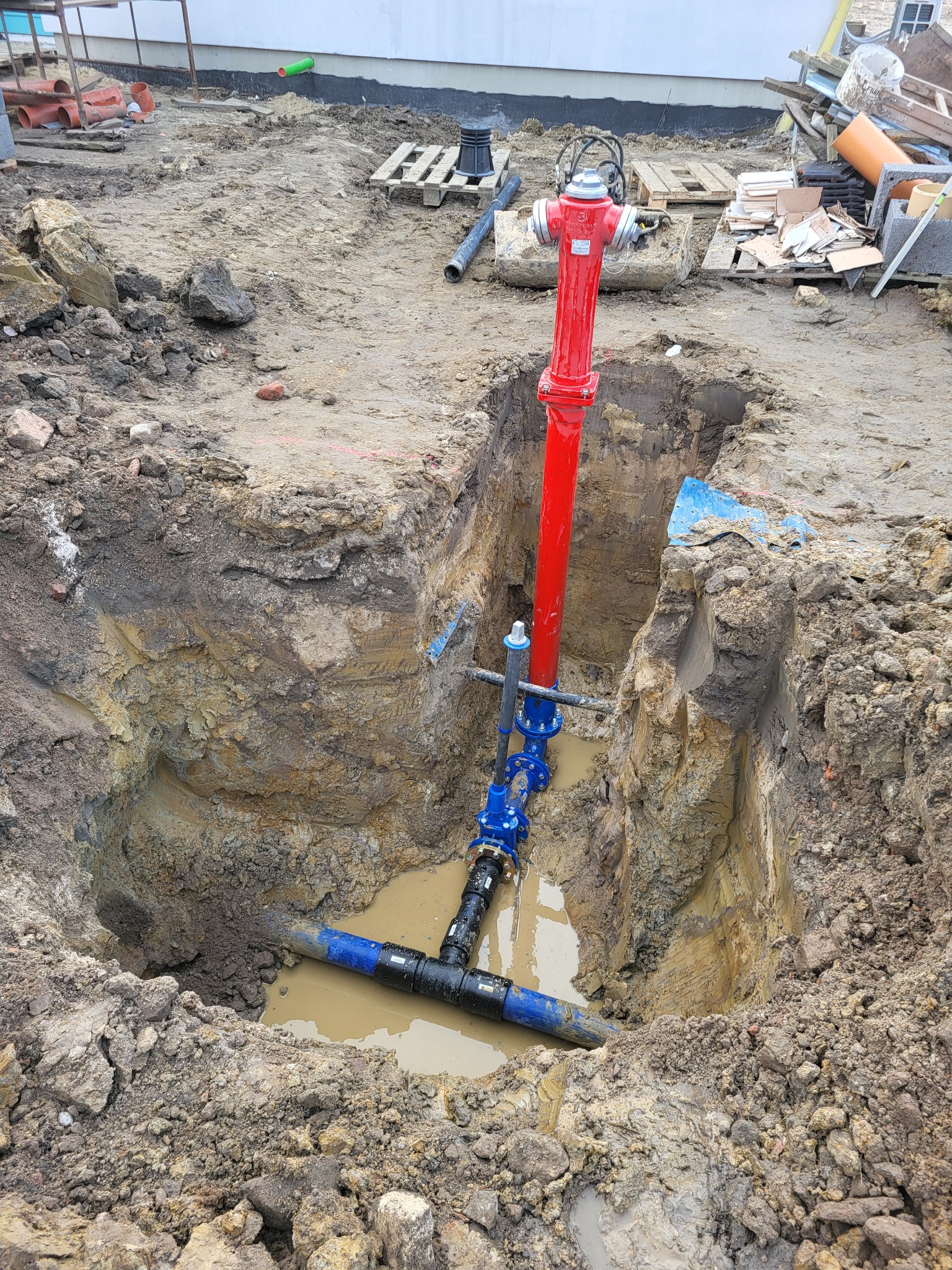 Czerwony hydrant w wykopie z instalacją wodociągową. Widoczne złącza i rury w ziemi. Prace ziemne w toku, teren budowy.