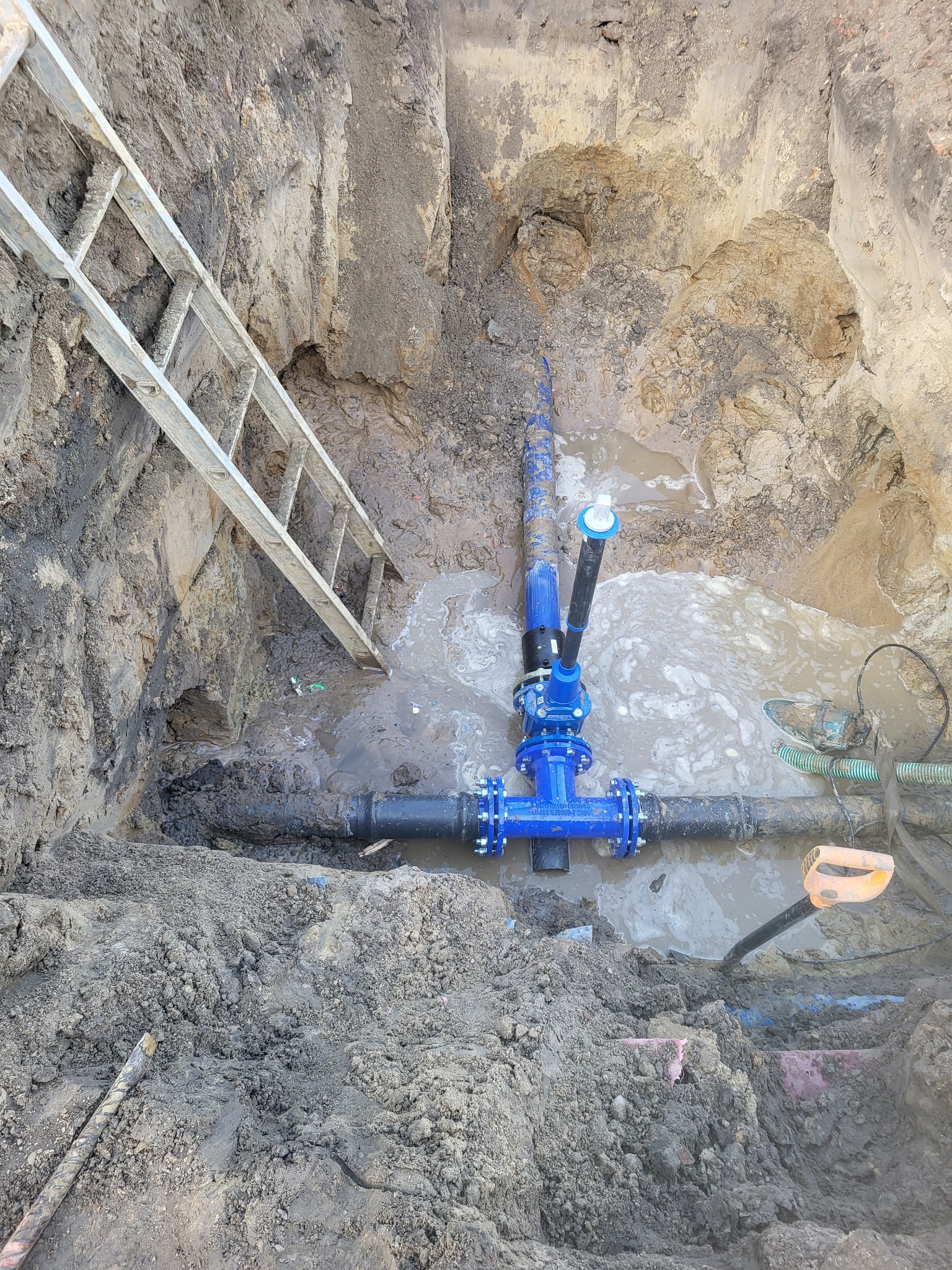 Wykop z wodą, w nim rury wodociągowe z niebieskimi złączkami. Ziemia w wykopie mokra, drabina oparta o ścianę wykopu. Widoczne narzędzia.