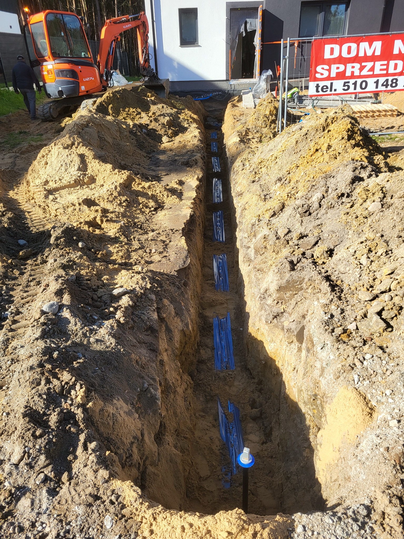 Wykop pod instalację z widocznymi niebieskimi rurami w ziemi. W tle koparka i budynek z banerem reklamowym. Ujęcie z góry, oświetlenie słoneczne.