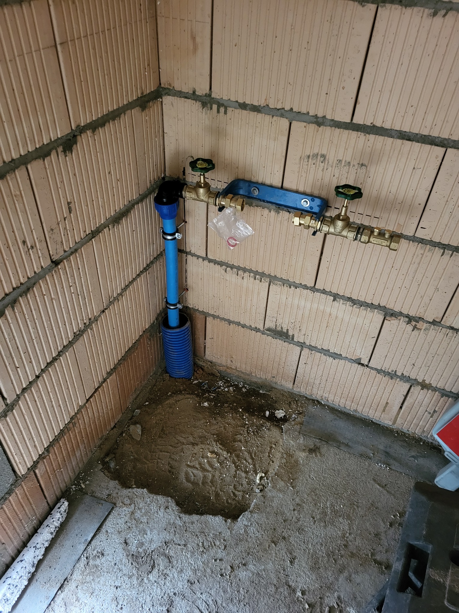 Niebieska rura instalacji wodnej z zaworami zamontowana przy ścianie z pustaków ceramicznych. Widoczne elementy mocujące i fragment posadzki.