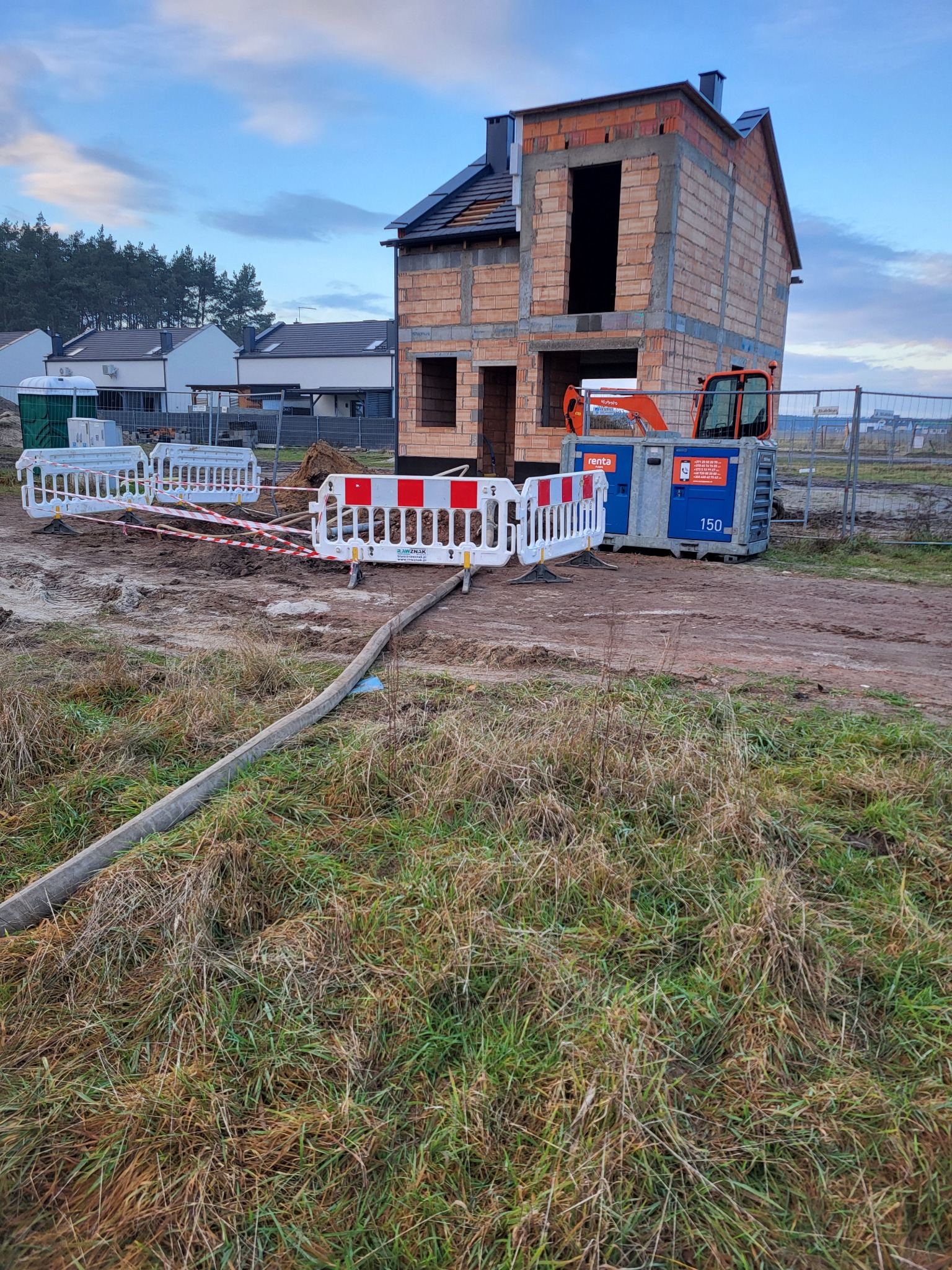 Budowa domu z pustaków. Na pierwszym planie ogrodzenie budowy, pompa do betonu i trawa. W tle inne domy i las. Widoczne niebo z chmurami.