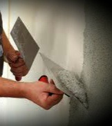 Nakładanie szpachli na ścianę za pomocą dwóch szpachelek, widoczne ręce pracownika i fragment ściany z nierówną powierzchnią, przygotowywanej do dalszych prac wykończeniowych.