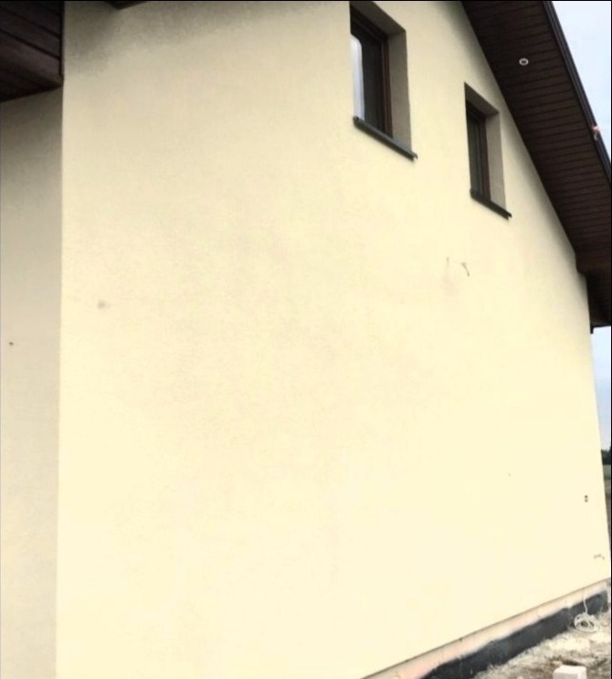 Ściana zewnętrzna domu jednorodzinnego po nałożeniu jasnobeżowego tynku elewacyjnego, widoczne dwa okna z ciemnymi ramami, brązowe podbitki dachowe i fundament zabezpieczony czarną izolacją.