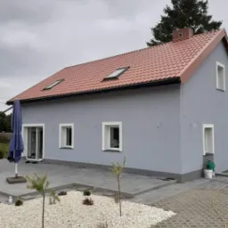 Szary dom jednorodzinny z czerwoną dachówką, dwoma oknami dachowymi i nową elewacją, widok z boku na ogród z białym żwirem i młodymi drzewkami.