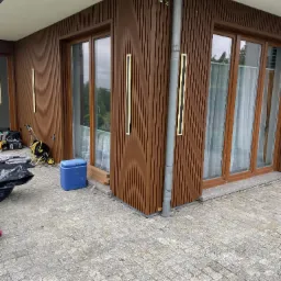 Wykończenie elewacji budynku z brązowymi panelami pionowymi, oświetleniem LED i drewnianymi oknami. Widoczne narzędzia i materiały budowlane.