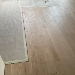 Układanie jasnych paneli podłogowych w pokoju, widoczny fragment podłogi zabezpieczony folią i taśmą malarską, w tle wiadro z narzędziami malarskimi.