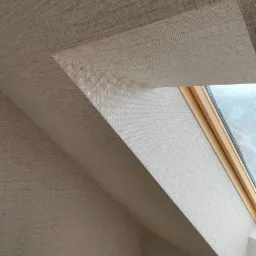 Beżowa tapeta o delikatnej strukturze na skośnej ścianie poddasza, z widocznym fragmentem okna dachowego z drewnianą ramą.