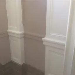 Pomieszczenie z pomalowanymi ścianami na jasny róż, z białymi, dekoracyjnymi panelami ściennymi i kolumnami, oraz szarą podłogą.