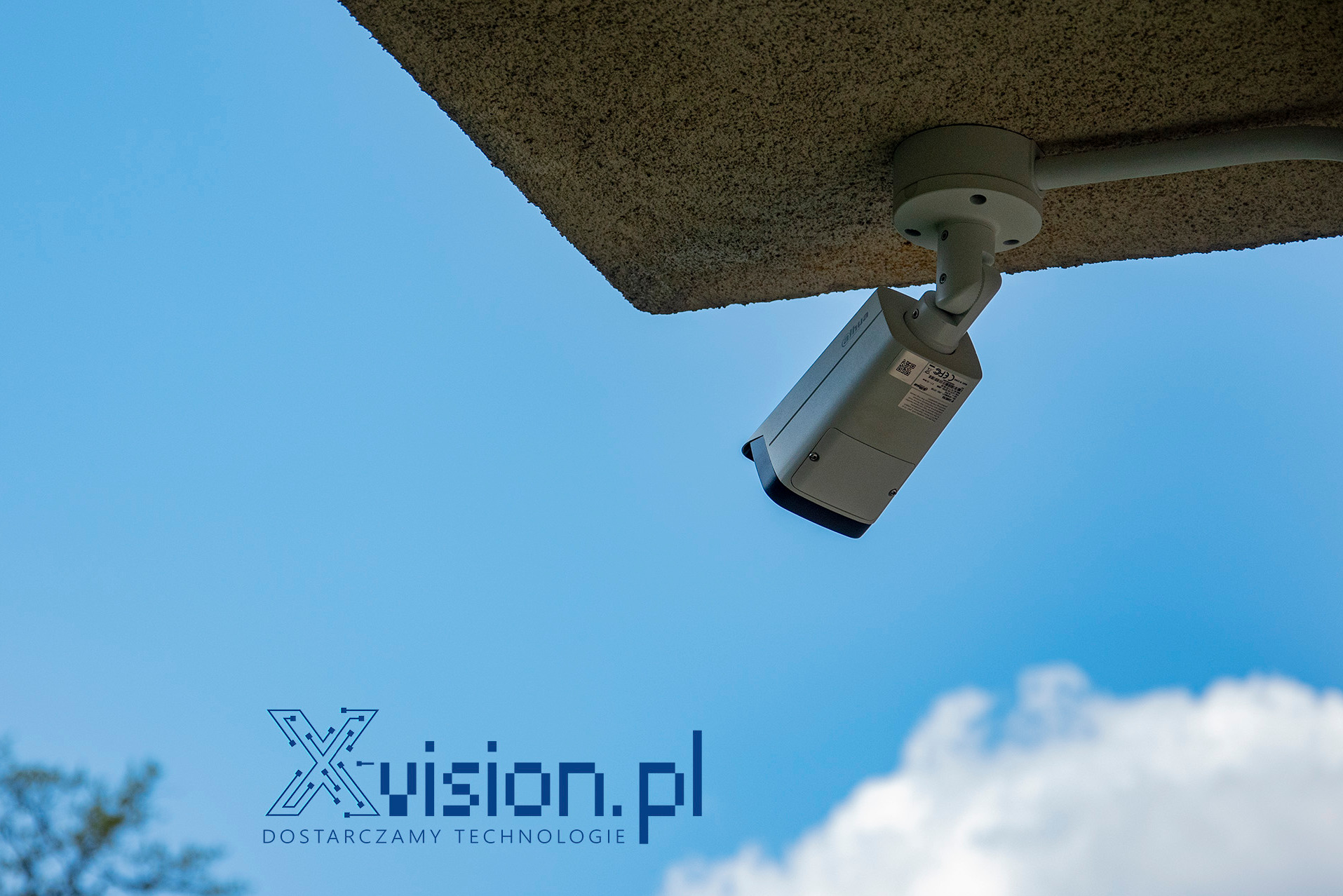 Kamera 8Mpix (4K) marki Dahua zamontowana pod wystającym fragmentem elewacji budynku w Suchej Beskidzkiej, widok w górę na błękitne niebo z chmurami.