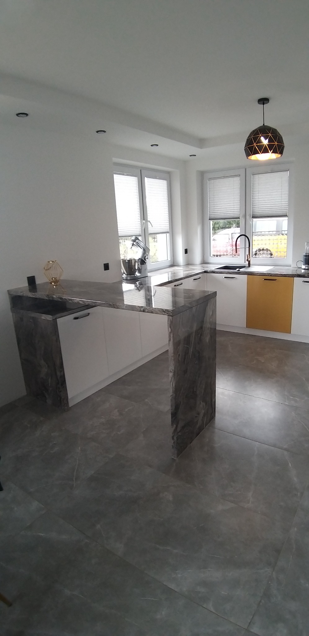 Nowoczesna kuchnia z wyspą wykończoną blatem z szarego marmuru, białe szafki, złote akcenty i geometryczna lampa wisząca.