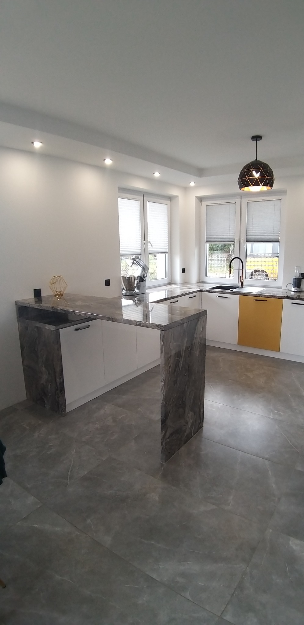 Nowoczesna kuchnia z wyspą wykończoną blatem imitującym marmur, białe szafki z czarnymi uchwytami, żółty front szafki, szare płytki podłogowe, okna z białymi żaluzjami, czarna bateria i ozdobna lampa.