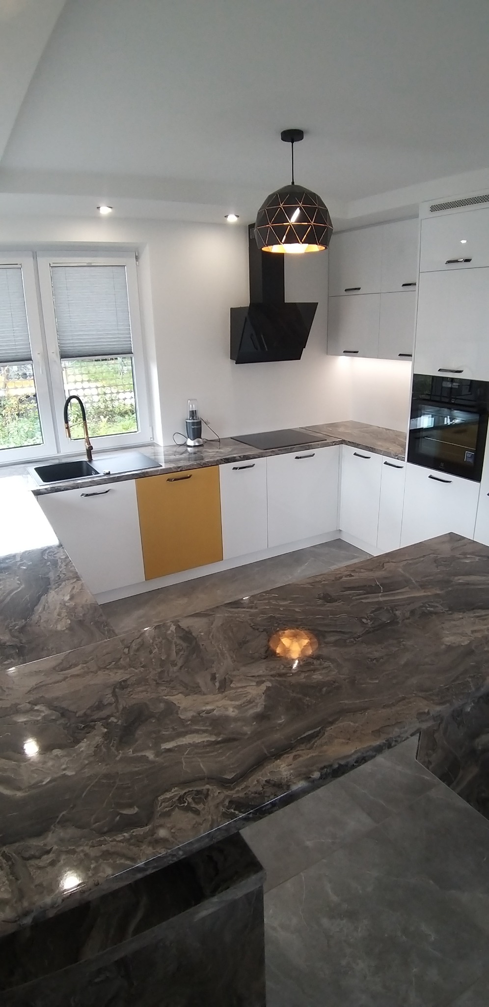 Nowoczesna kuchnia z białymi szafkami, blatem imitującym marmur, czarnym okapem i złotym akcentem na jednej z szafek.