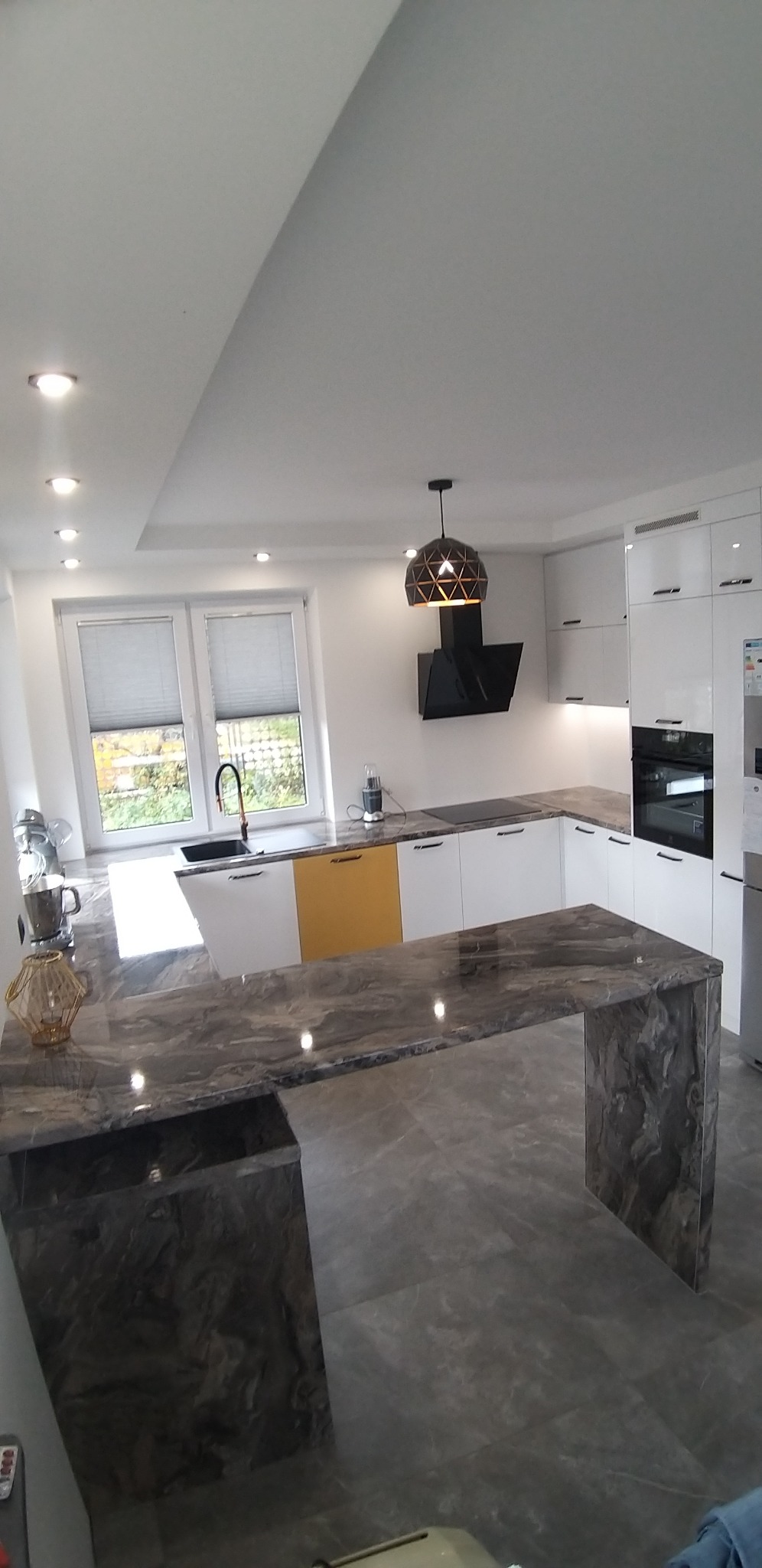 Nowoczesna kuchnia z białymi szafkami, blatem kuchennym z efektem marmuru i wyspą z elementami w kolorze musztardowym, oświetlona punktowymi lampami i designerską lampą wiszącą.