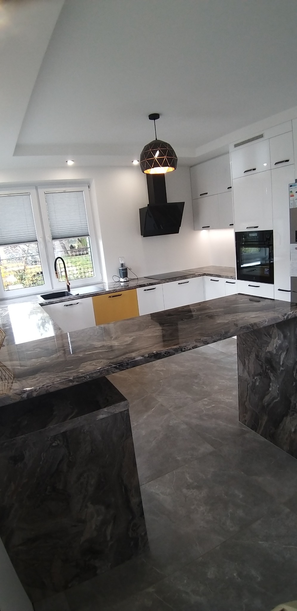 Nowoczesna kuchnia z białymi szafkami, marmurowym blatem i wyspą, czarnym okapem i geometryczną lampą wiszącą.