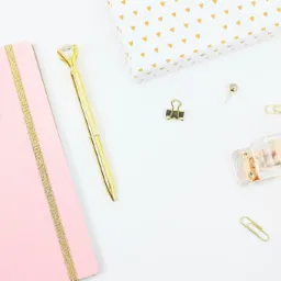 Różowy notatnik ze złotą gumką i klipsem, złoty długopis z kryształkiem, pudełko w trójkąty, spinacze, pinezka i taśma klejąca na białym tle.