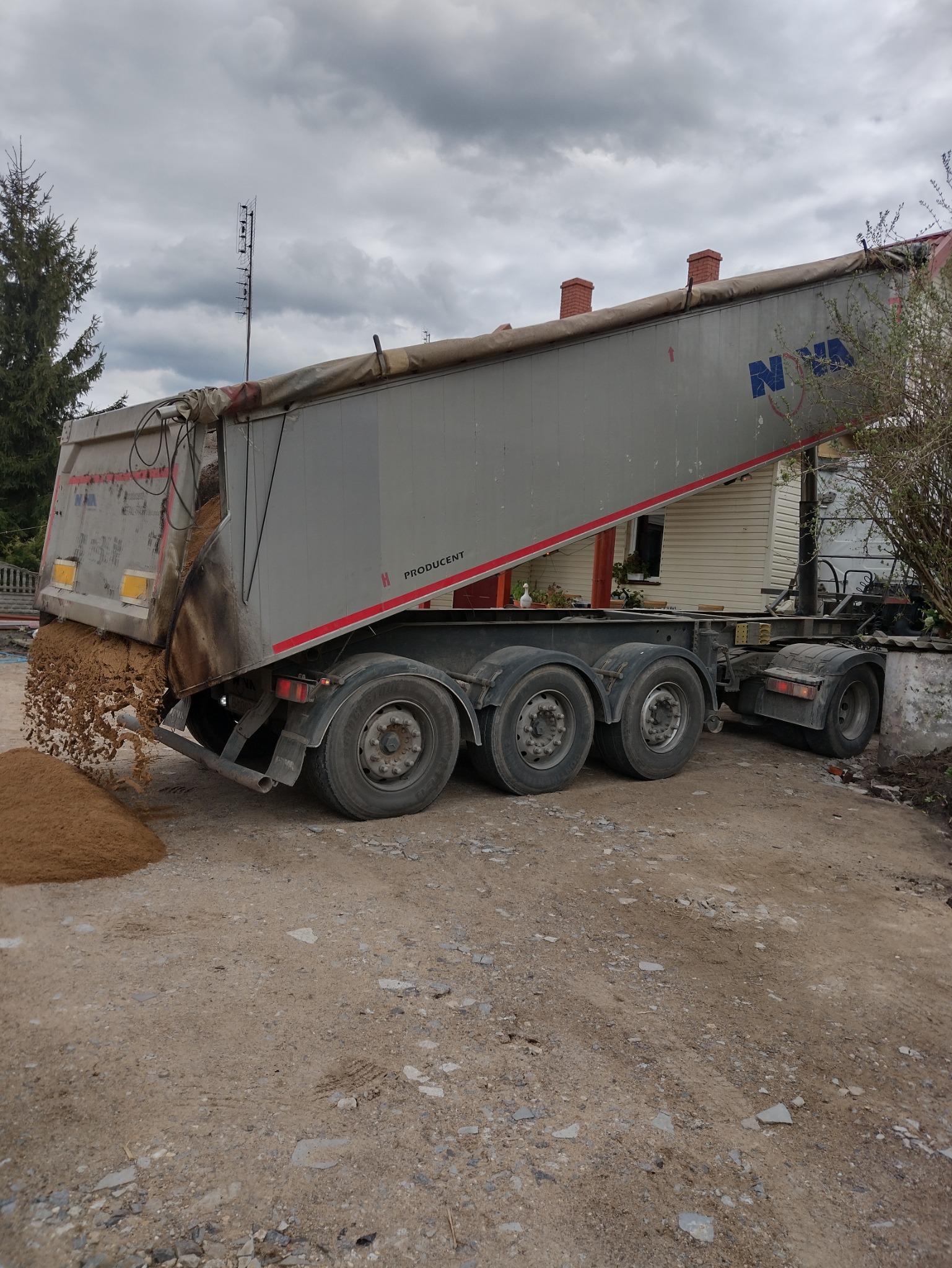 Ciężarówka z naczepą wyładowuje piasek na nierównym terenie przed domem z ceglanymi kominami, widoczne błotniste ślady opon.