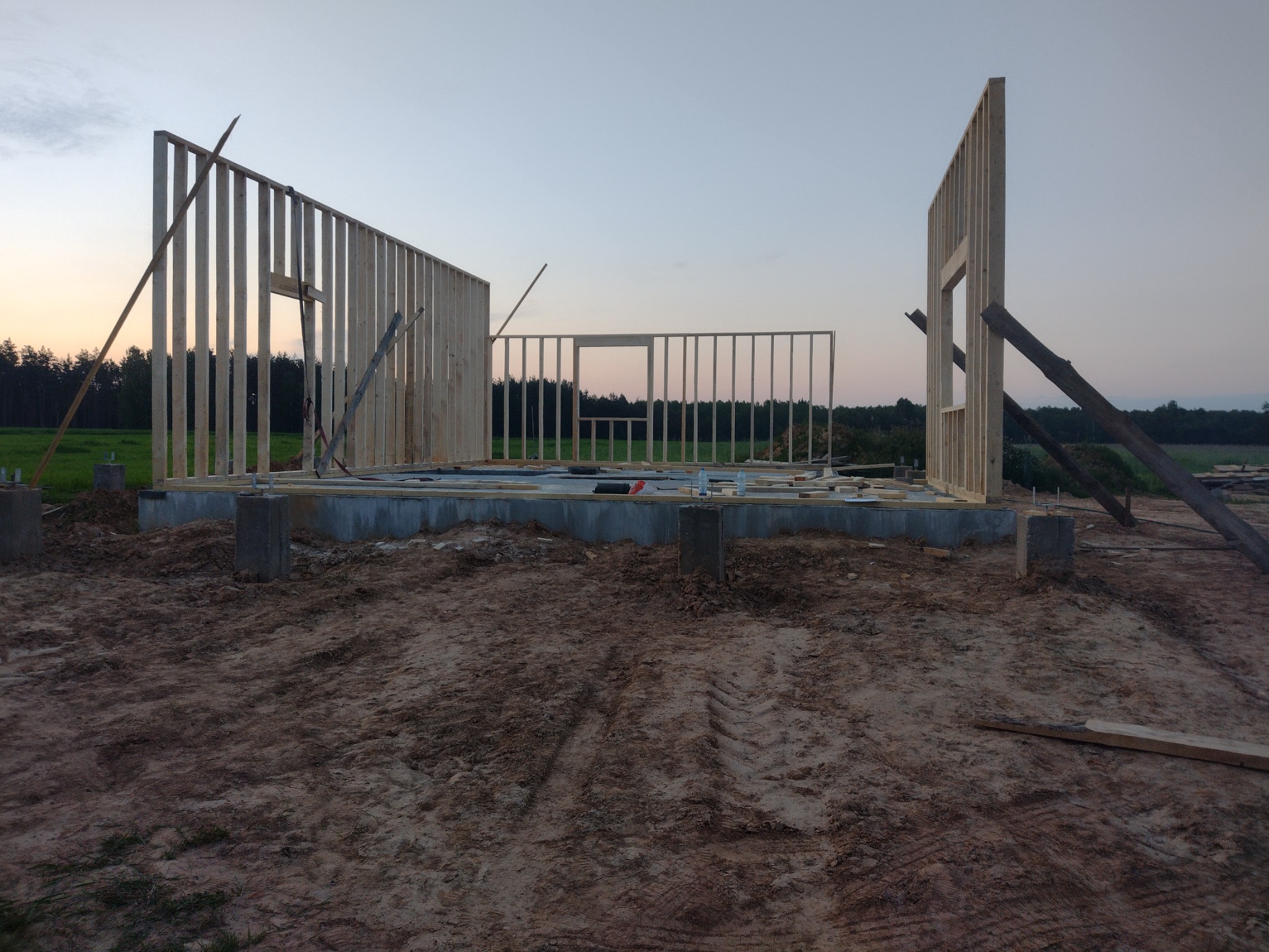 Wznoszenie konstrukcji szkieletowej domu: widok na fundament z betonowymi słupami i częściowo wzniesione ściany z drewnianych belek na tle zachodzącego słońca.