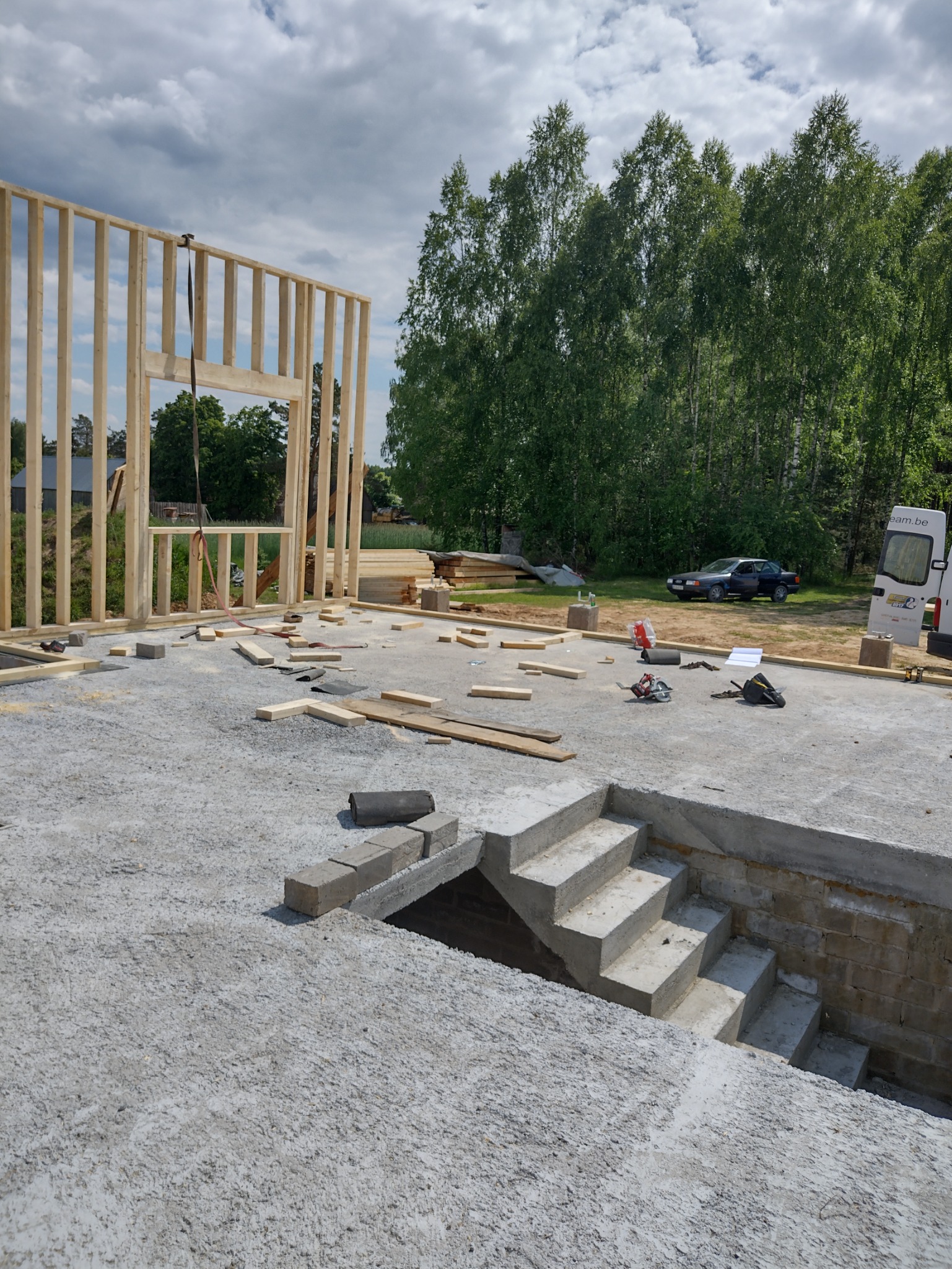 Wznoszenie drewnianej konstrukcji szkieletowej na betonowej płycie fundamentowej z widocznymi schodami do piwnicy, na tle zieleni i pochmurnego nieba.