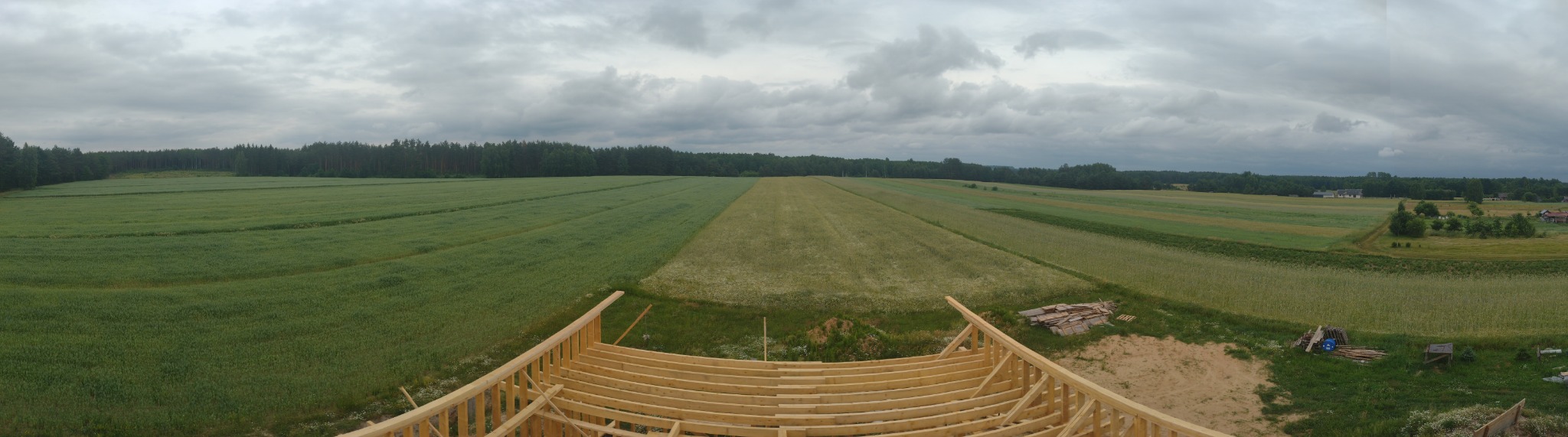 Panoramiczny widok z góry na drewnianą konstrukcję dachu w trakcie budowy, z rozległymi polami i lasem w tle pod pochmurnym niebem.