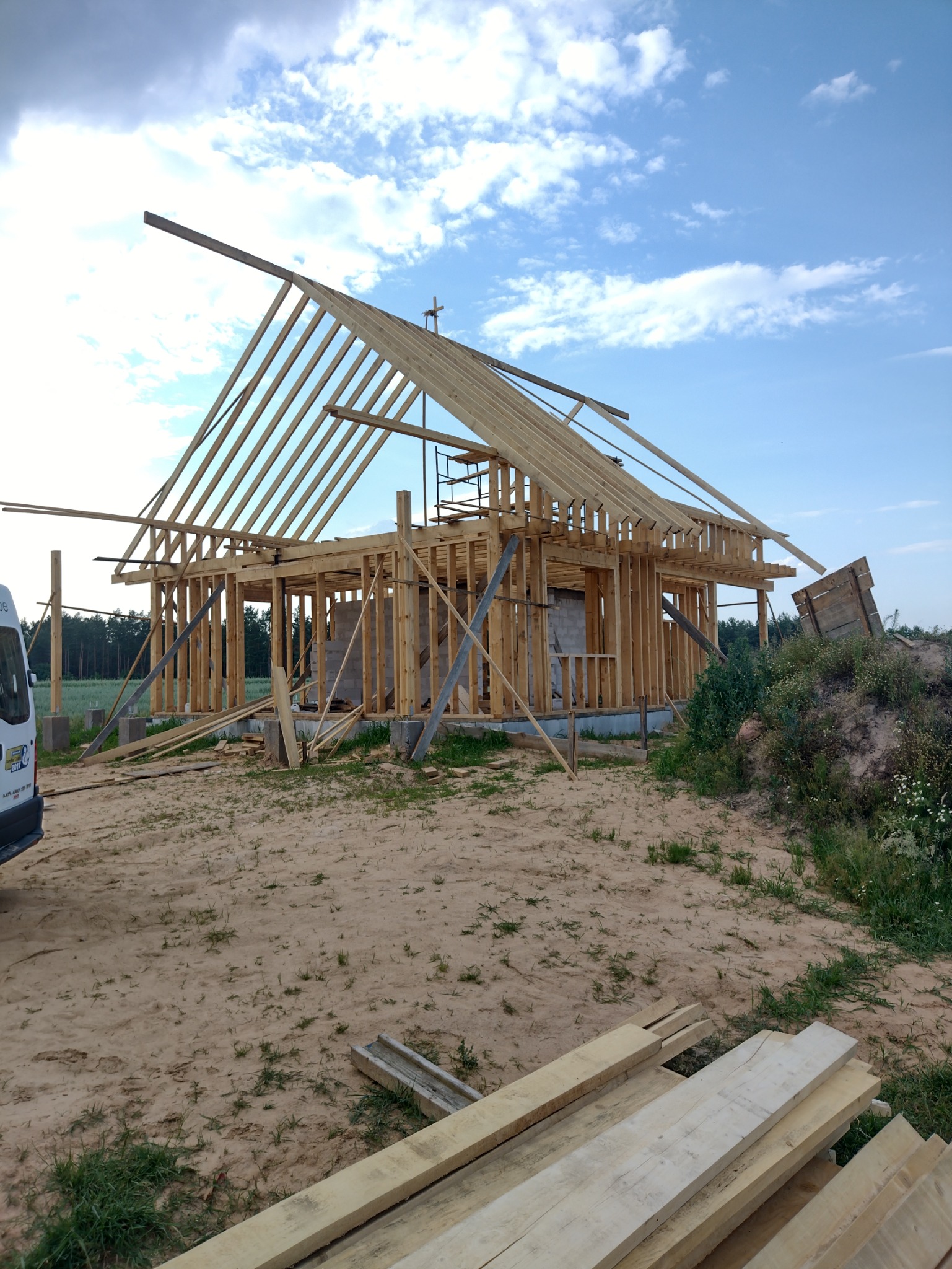 Wznoszenie konstrukcji drewnianej domu jednorodzinnego z widocznymi elementami więźby dachowej i ścian szkieletowych na tle błękitnego nieba.