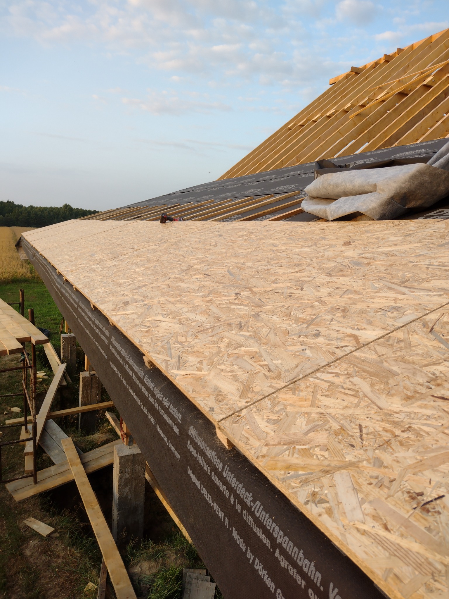 Konstrukcja dachu w trakcie budowy: widoczne drewniane krokwie, płyty OSB, czarna membrana z nadrukiem producenta oraz narzędzie do mocowania. W tle pole i drzewa.