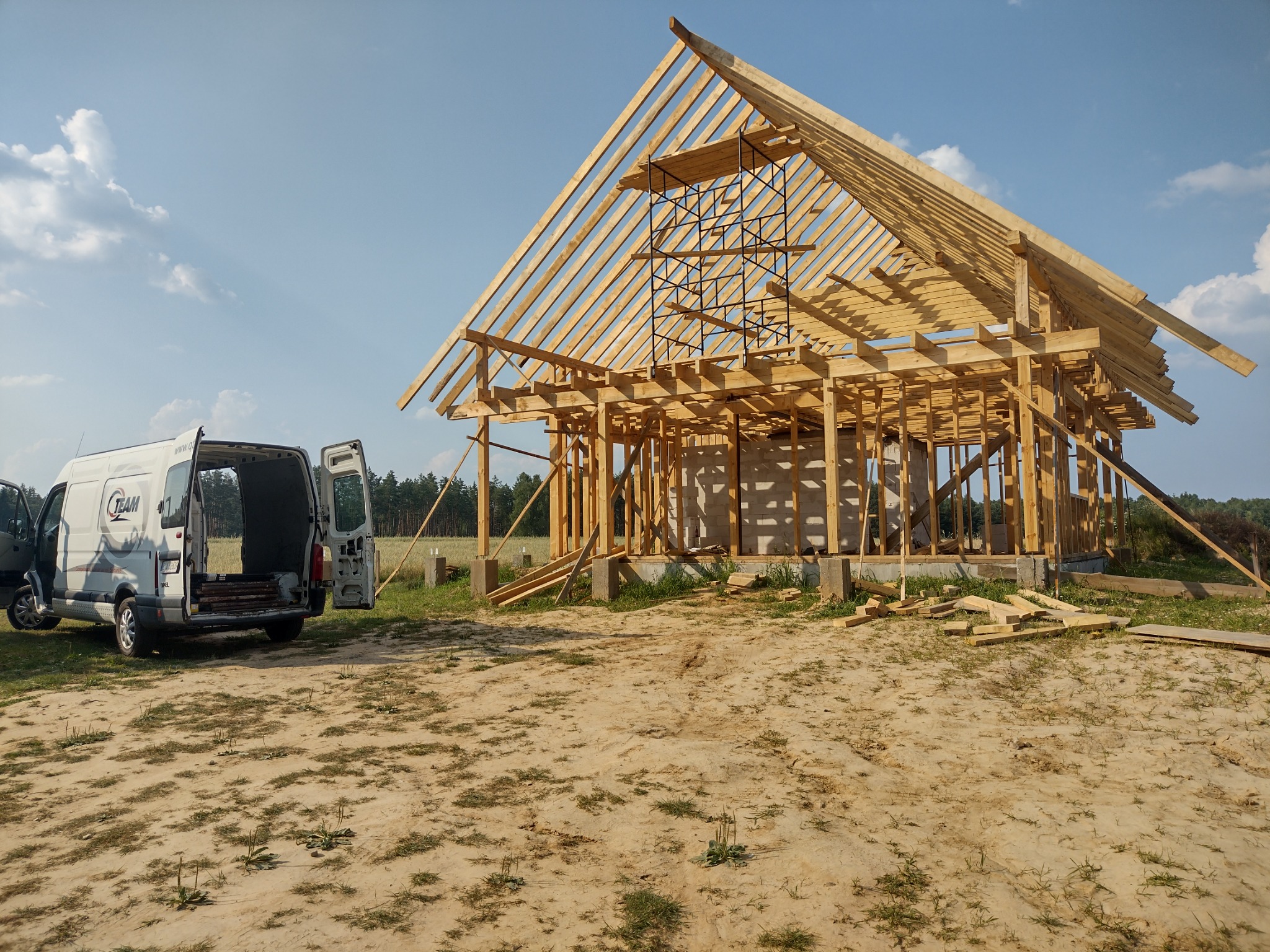 Konstrukcja drewniana domu w trakcie budowy, widoczny szkielet dachu i ścian, na pierwszym planie samochód dostawczy z otwartymi drzwiami, piaszczyste podłoże.