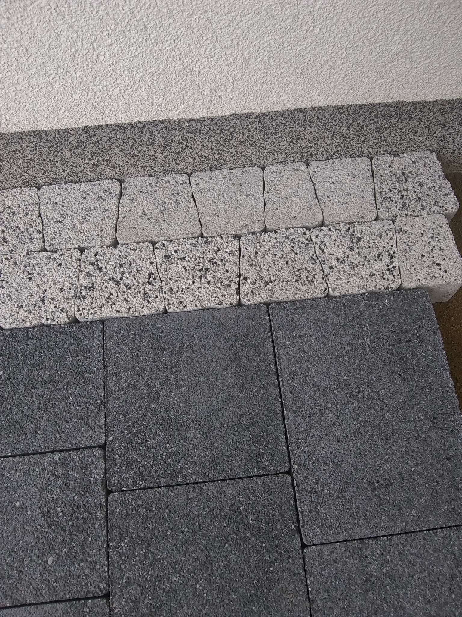Ułożone obok siebie kostki brukowe w odcieniach szarości, z widoczną fakturą i krawędziami, na tle ściany o zróżnicowanej strukturze.