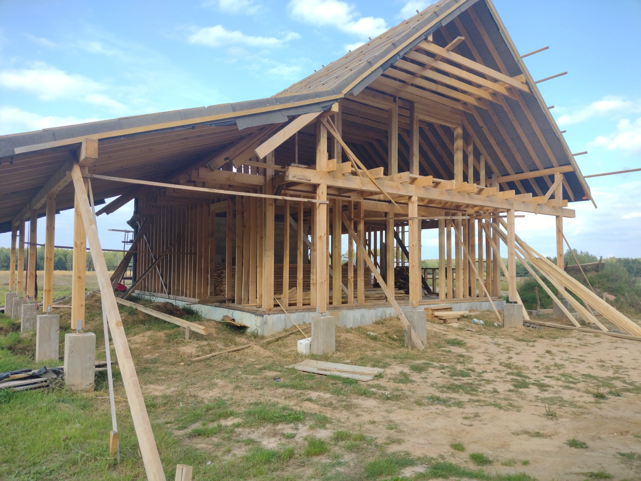 Wznoszenie konstrukcji domu z drewnianych belek na betonowym fundamencie, widoczne elementy dachu i ścian, teren budowy z trawą i niebem w tle.