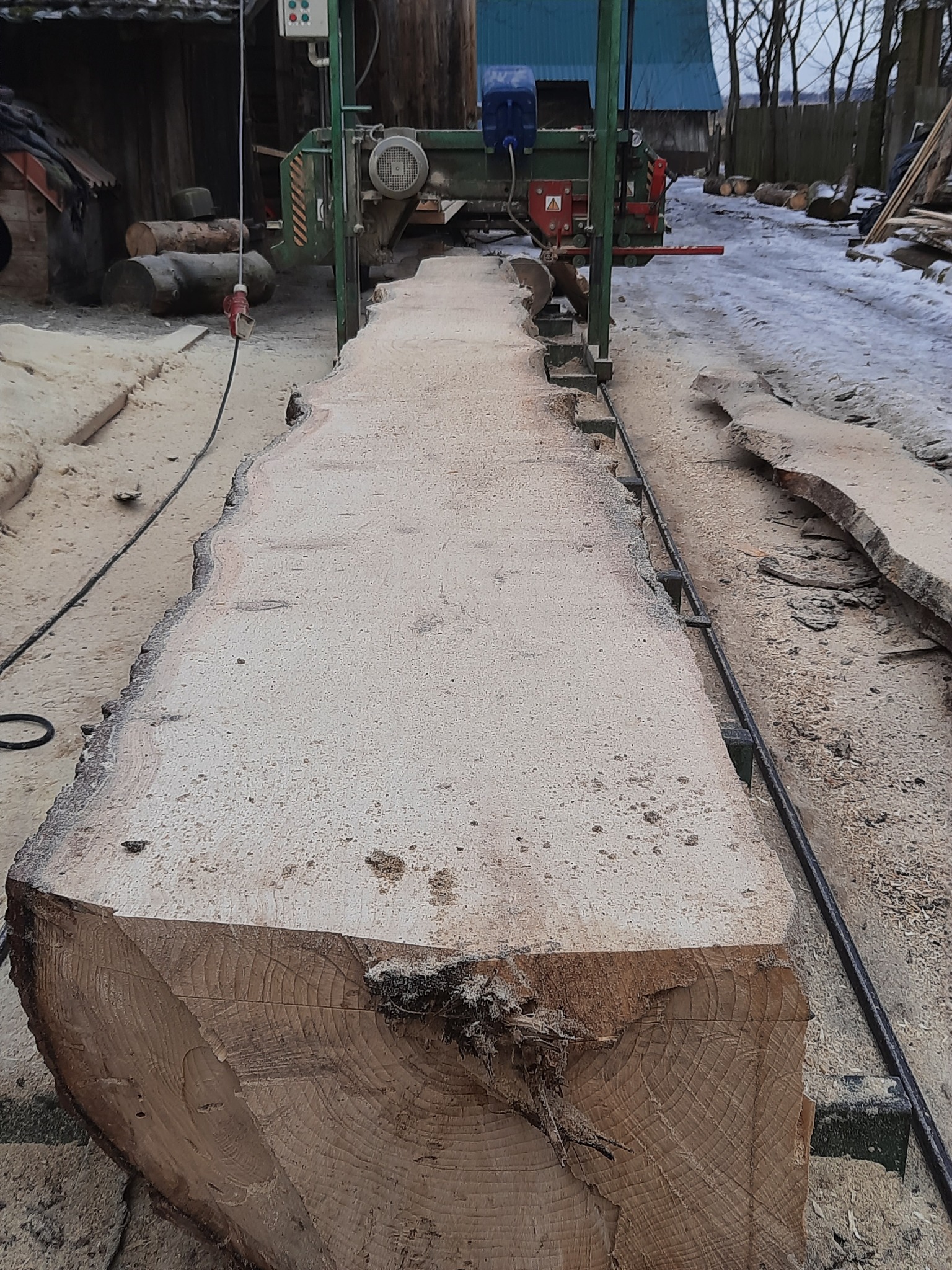 Przecieranie kłody drewna na traku taśmowym, widoczne świeże deski i trociny, w tle budynek gospodarczy i ośnieżony teren.