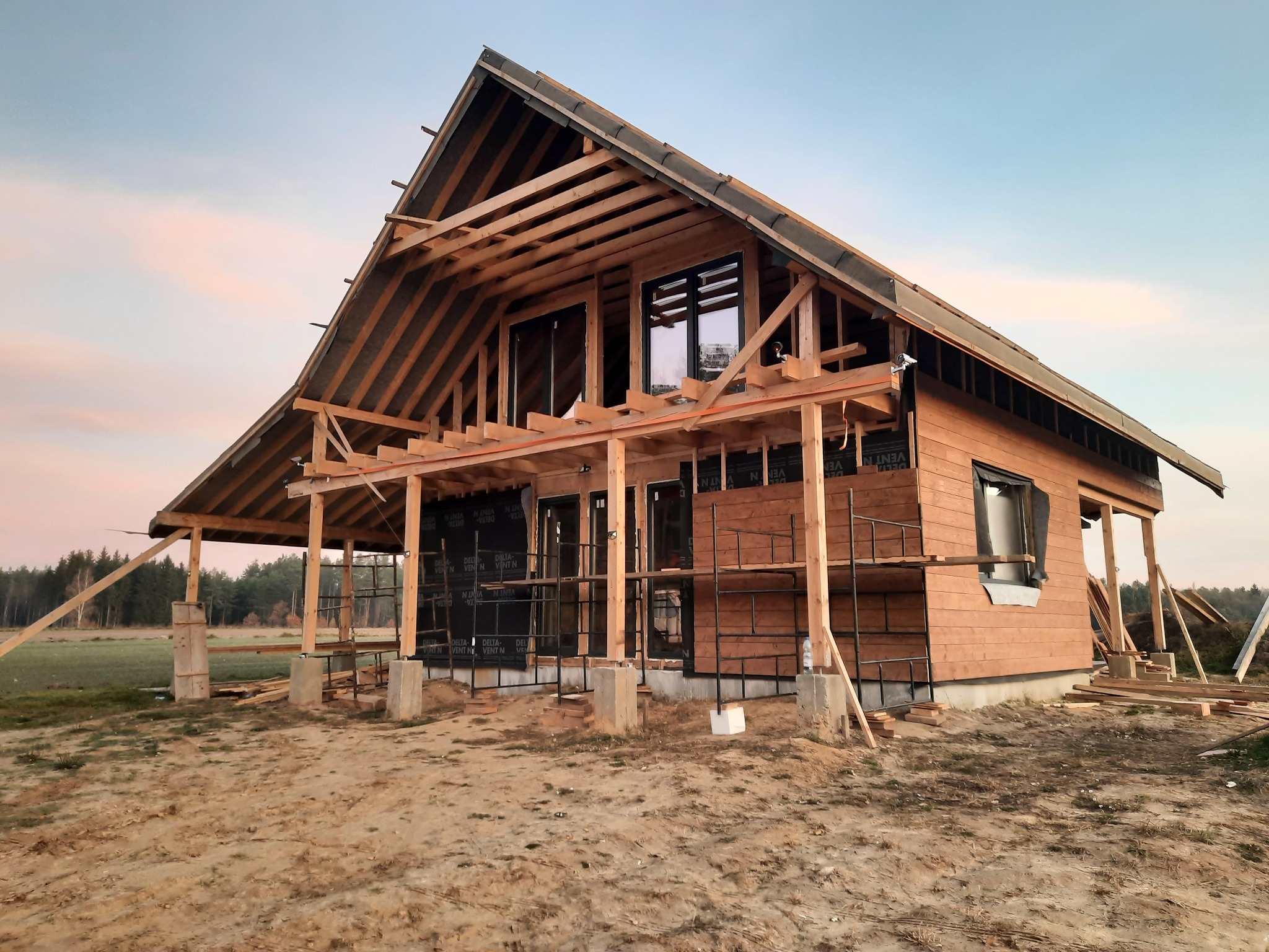 Budowa domu z drewnianą konstrukcją szkieletową, widoczne elementy dachu i ściany pokryte deskami, rusztowania dookoła, teren budowy.