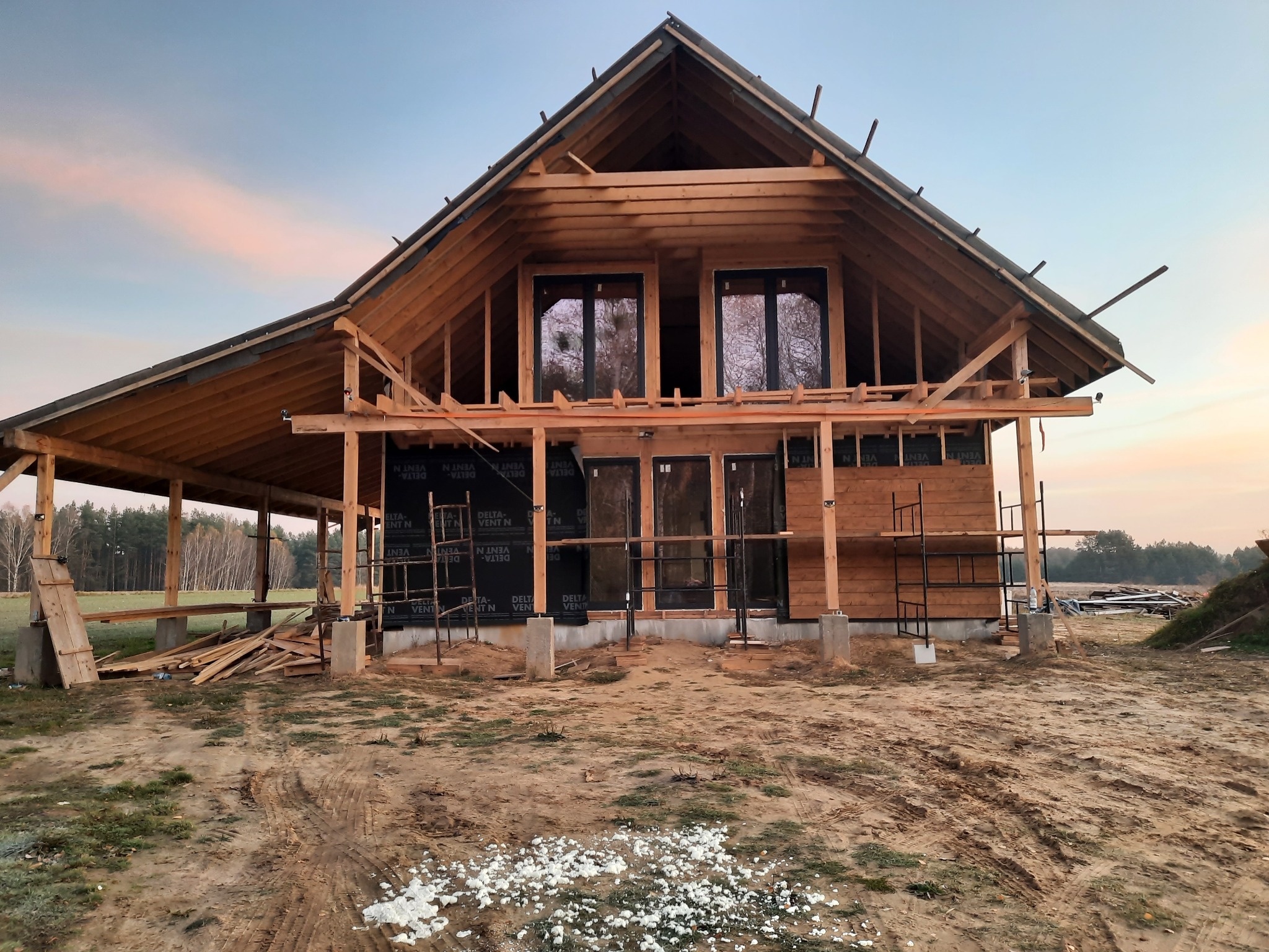 Surowy stan budowy domu szkieletowego w Białymstoku, widoczne drewniane elementy konstrukcyjne, czarna folia izolacyjna, rusztowania i teren budowy.