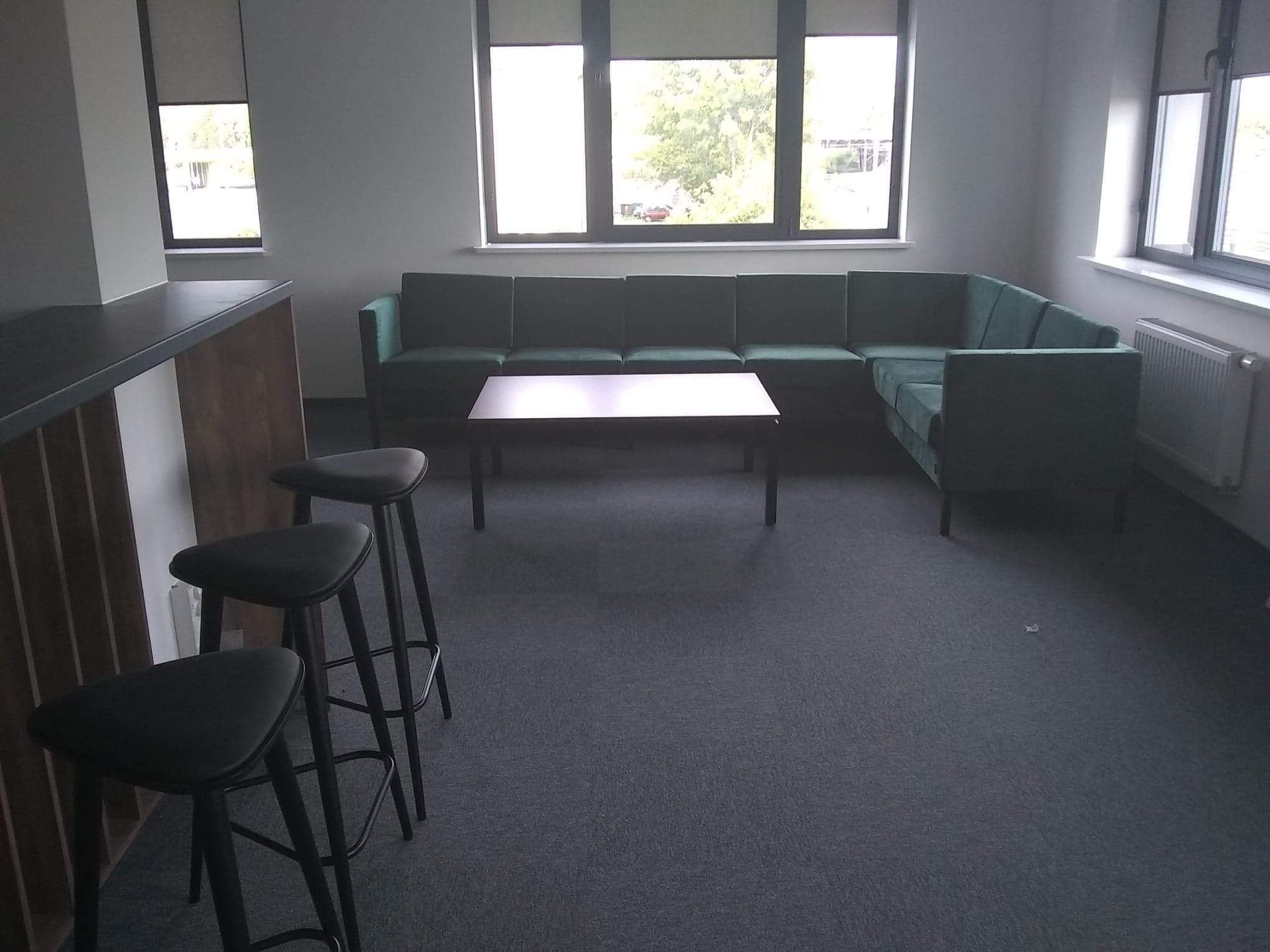 Wnętrze biura z zielonym narożnikiem, stolikiem kawowym i trzema czarnymi stołkami barowymi przy zabudowie kuchennej, szara wykładzina dywanowa.