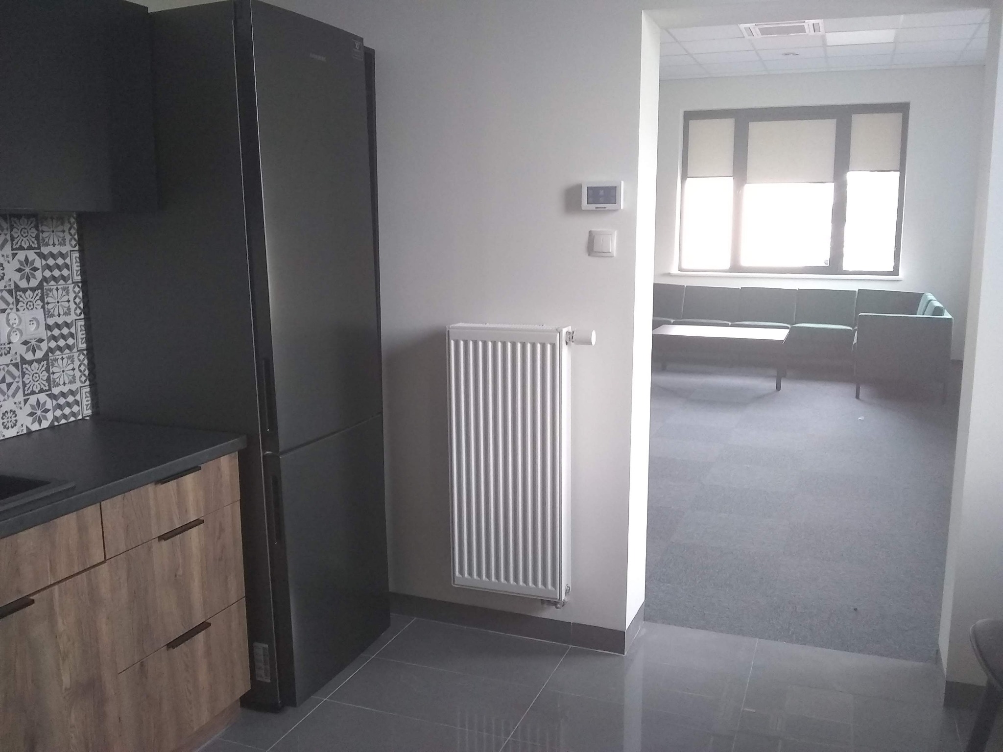 Nowoczesne wnętrze biurowe z aneksem kuchennym, szarą podłogą, grzejnikiem i widokiem na strefę relaksu z zieloną sofą.