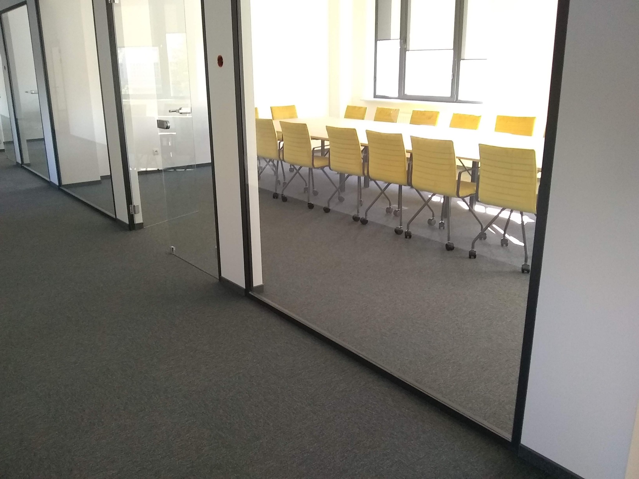 Nowoczesne biuro z przeszklonymi ścianami działowymi, szarym dywanem i salą konferencyjną z żółtymi krzesłami w tle.