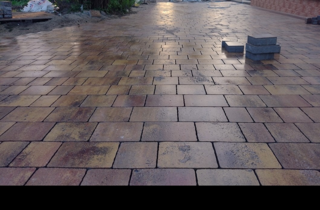 Ułożona nawierzchnia z kostki brukowej w odcieniach brązu i beżu, z widocznymi śladami wilgoci, z kilkoma nieułożonymi kostkami w górnym prawym rogu oraz przygotowanym podłożem w tle.