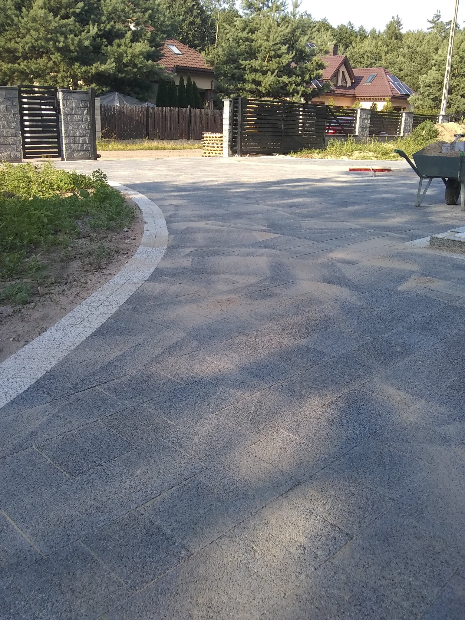 Ułożona kostka brukowa na podjeździe w Białymstoku, widoczna brama wjazdowa i dom w tle, taczka z piaskiem obok.