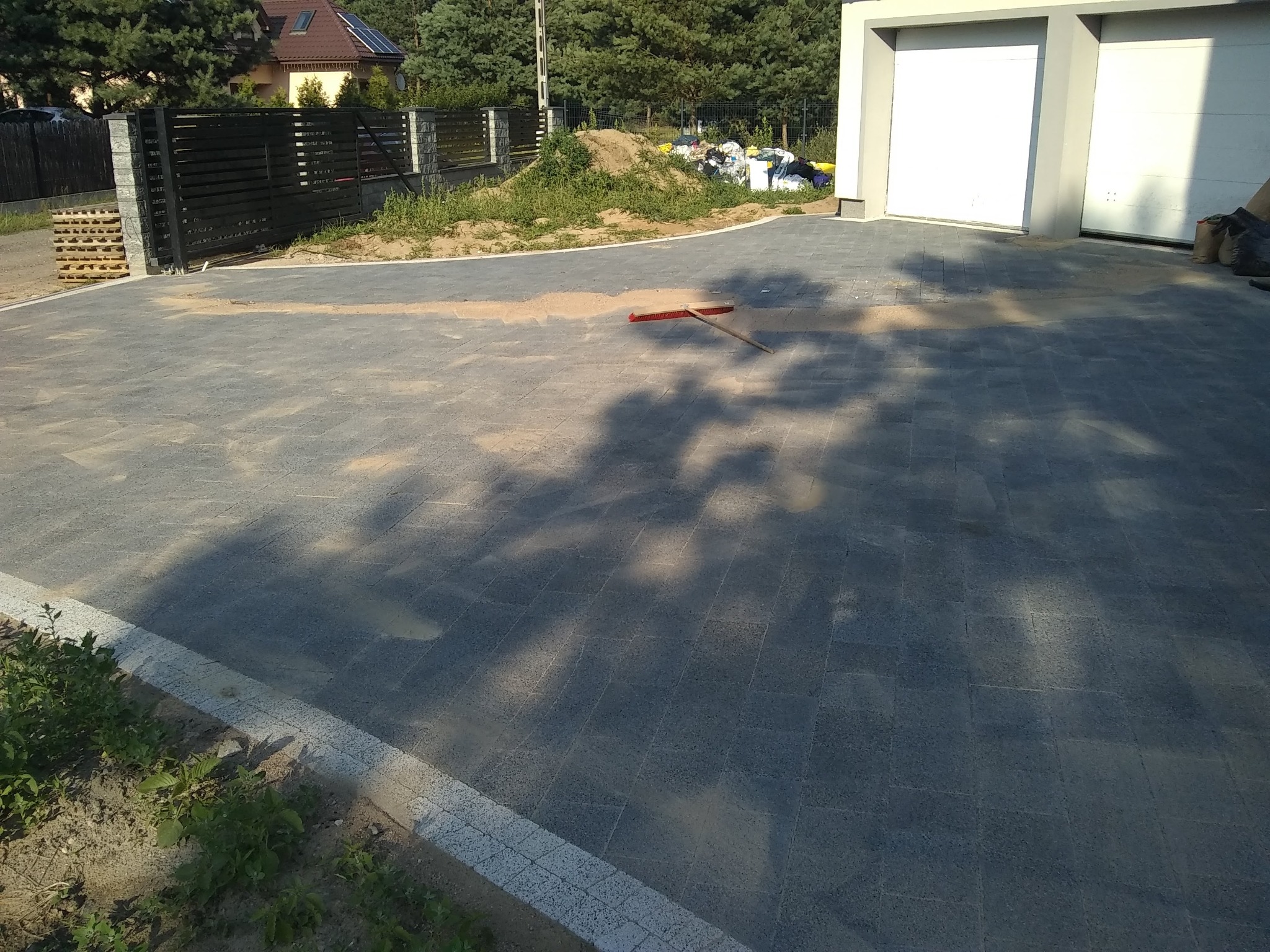 Ułożona szara kostka brukowa na podjeździe do garażu, z widoczną czerwoną miotłą i fragmentem ogrodzenia oraz stertą śmieci w tle.
