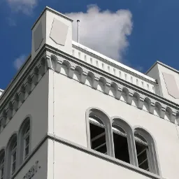 Biała wieża z oknami łukowymi i dekoracyjnymi elementami architektonicznymi na tle błękitnego nieba z chmurami, widziana z dołu.