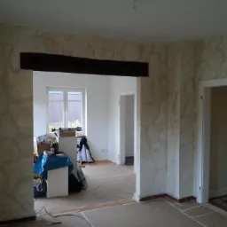 Pokój w trakcie prac wykończeniowych: ściany z dekoracyjną fakturą imitującą kamień, widoczny fragment drewnianej belki stropowej, podłoga zabezpieczona folią malarską, widoczne drzwi wewnętrzne...