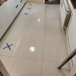 Białe płytki podłogowe w kuchni z zaznaczonymi miejscami niebieską taśmą, widoczny fragment białej zabudowy kuchennej i grzejnik.