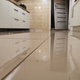 Daniel Przybylski - Błyszcząca, beżowa podłoga z płytek w łazience, widok z niskiej perspektywy na białe szafki kuchenne z drewnianym blatem, drzwi wejściowe i grzejnik.