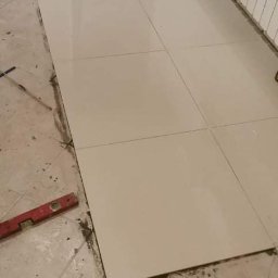 Daniel Przybylski - Ułożone jasne płytki podłogowe w trakcie prac wykończeniowych, widoczny poziomica, szpachelka i resztki zaprawy na starej podłodze.