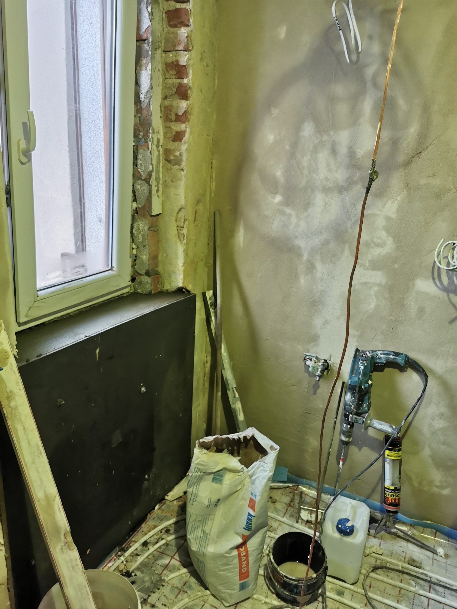 Łazienka w trakcie prac przygotowawczych: widoczne rury ogrzewania podłogowego, odsłonięta cegła przy oknie, wiadro z zaprawą, worek Knauf Band, wiertarka z mieszadłem i pistolet do pianki montażowej.