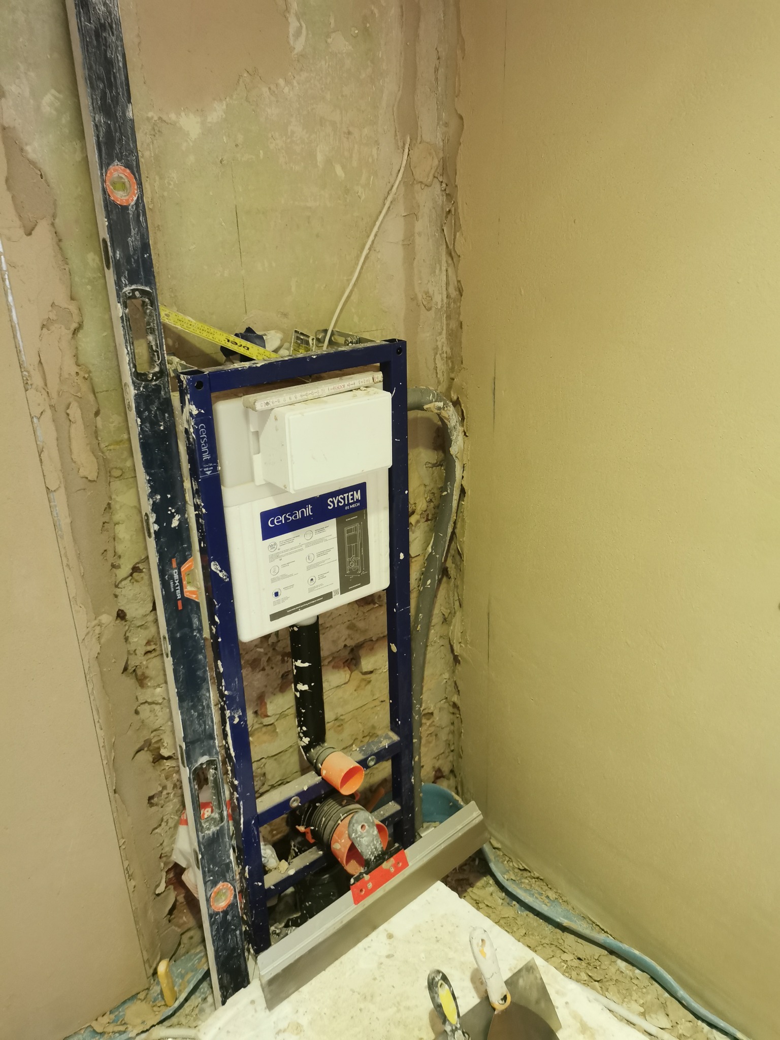 Instalacja podtynkowa WC Cersanit System w trakcie montażu, widoczny poziomica Dexter, nieotynkowana ściana z instalacjami i narzędzia na podłodze.