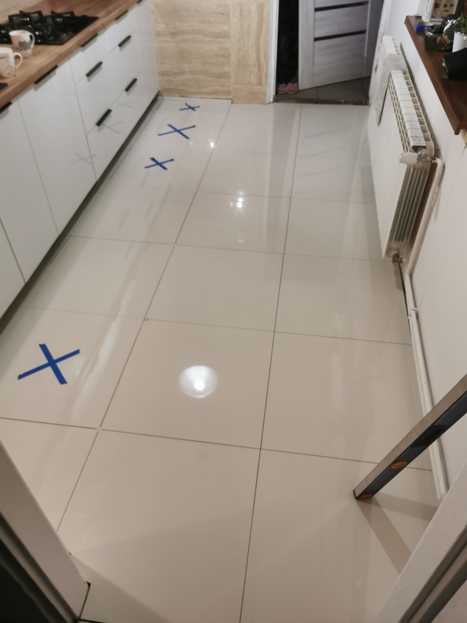 Białe płytki podłogowe w kuchni z zaznaczonymi miejscami niebieską taśmą, widoczny fragment białej zabudowy kuchennej i grzejnik.