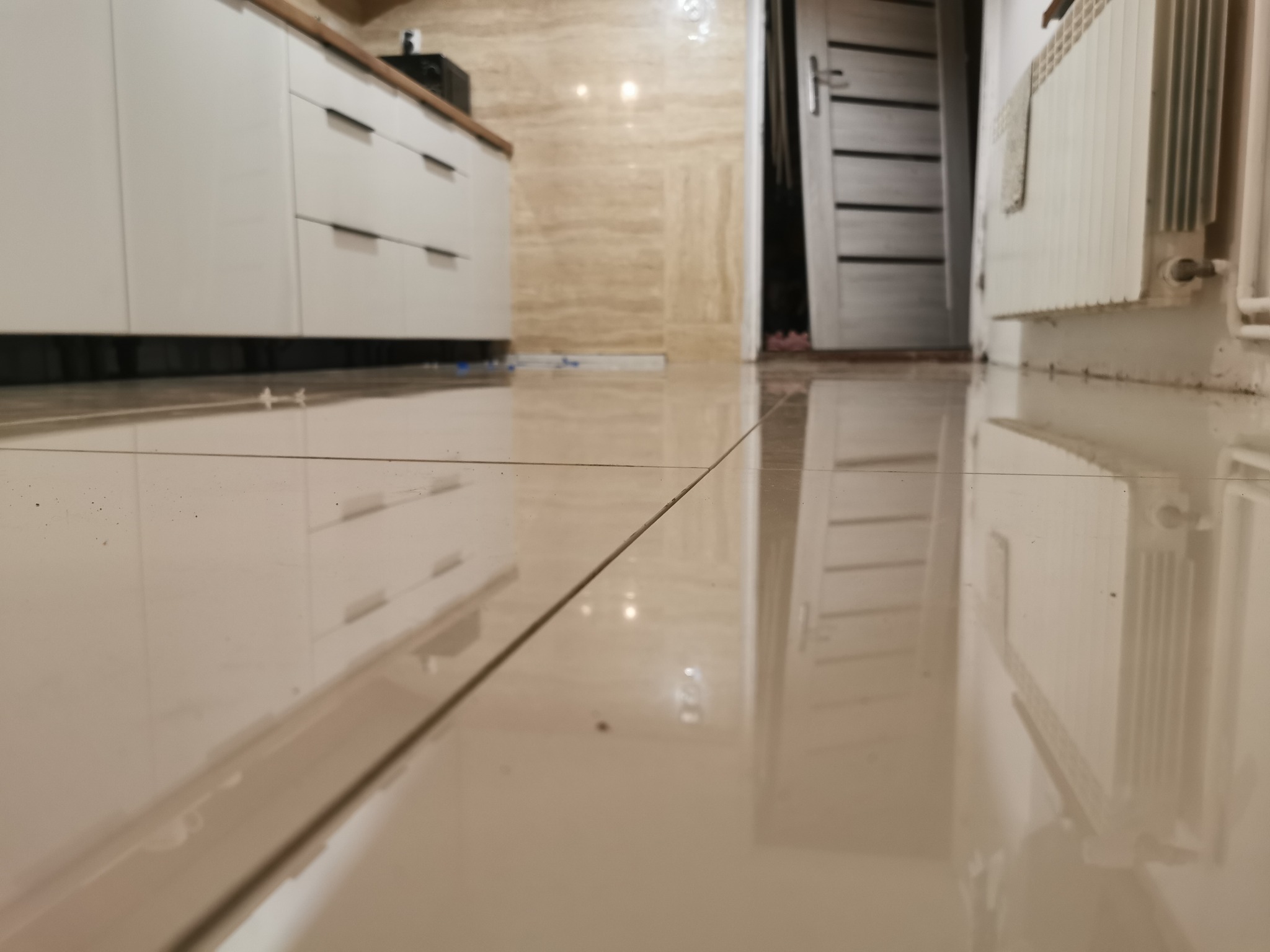 Błyszcząca, beżowa podłoga z płytek w łazience, widok z niskiej perspektywy na białe szafki kuchenne z drewnianym blatem, drzwi wejściowe i grzejnik.