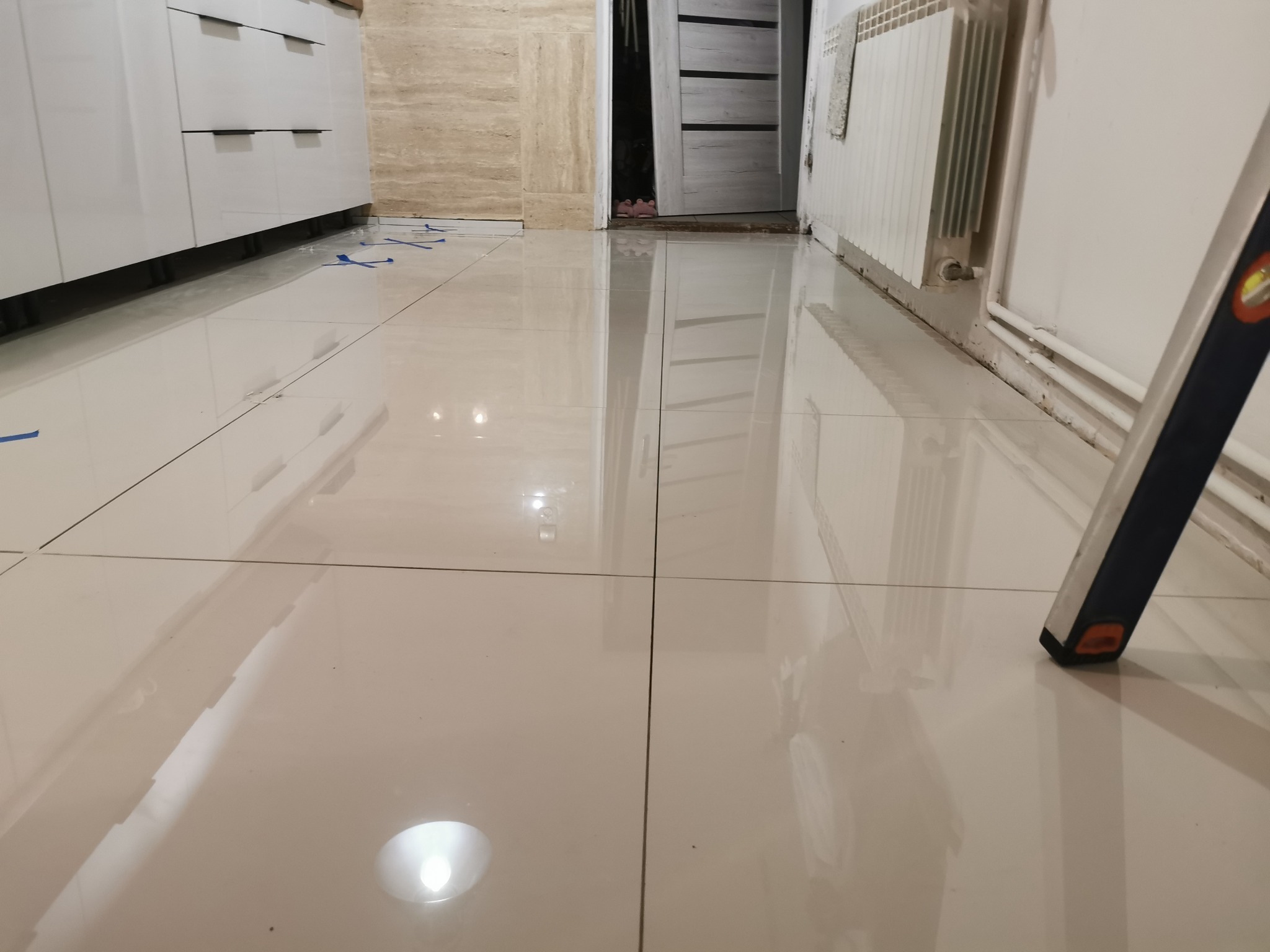 Błyszcząca, kremowa podłoga z płytek w trakcie układania, zabezpieczona niebieską taśmą, widoczny fragment poziomicy, białe meble i grzejnik na ścianie.